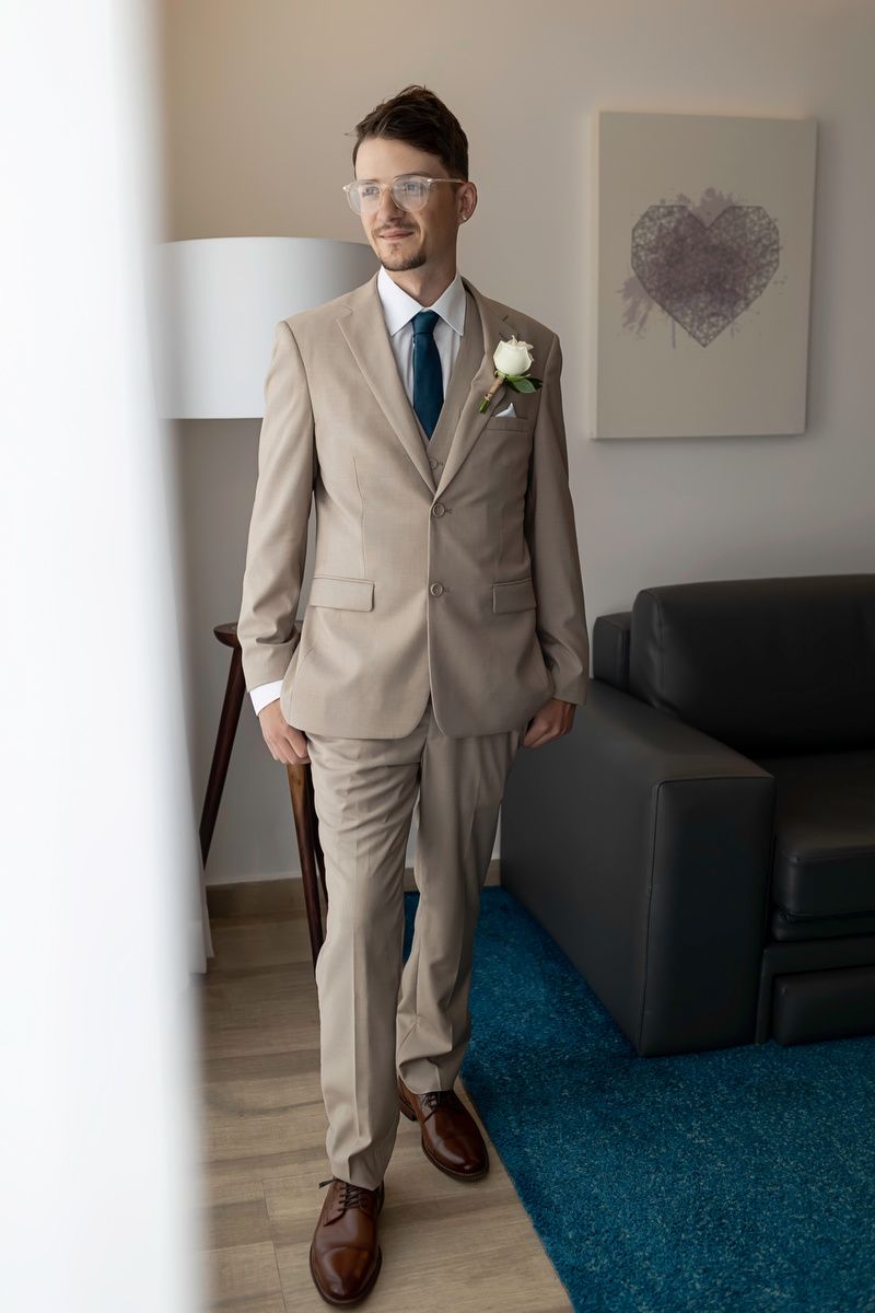 Groom in beige suit with boutonniere, navy tie, and glasses standing in modern room before wedding ceremony.