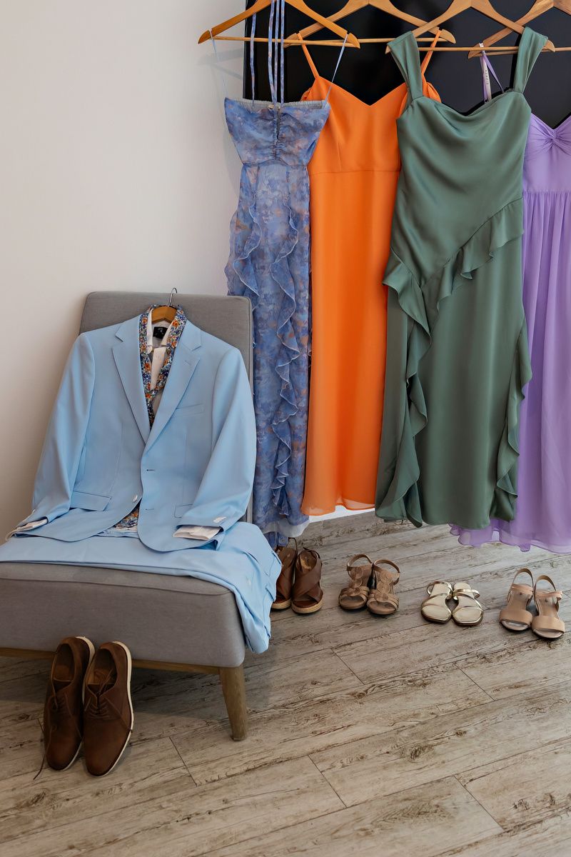 Light blue suit on chair beside colorful bridesmaid dresses in orange, green, and purple hanging on rack with shoes below.