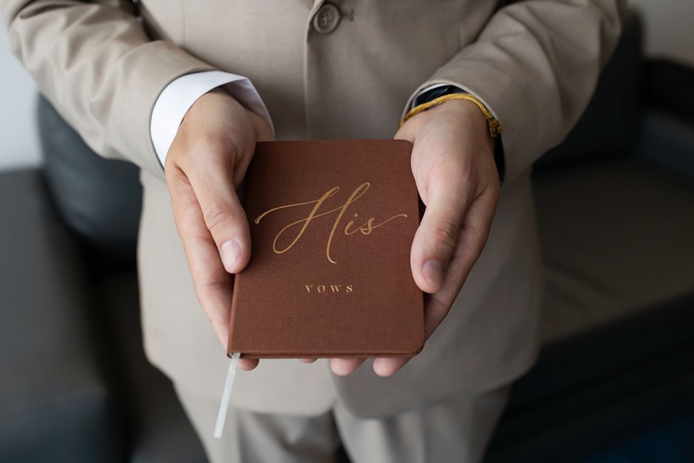 Groom holding a brown leather vow book with gold His script lettering during wedding ceremony.