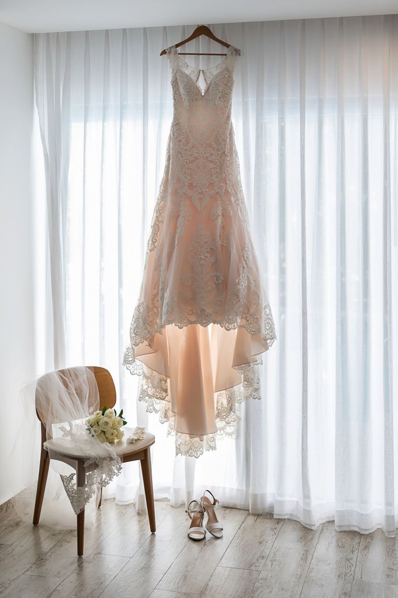 Elegant lace wedding dress hanging by window with bridal bouquet on chair and white heels on hardwood floor.