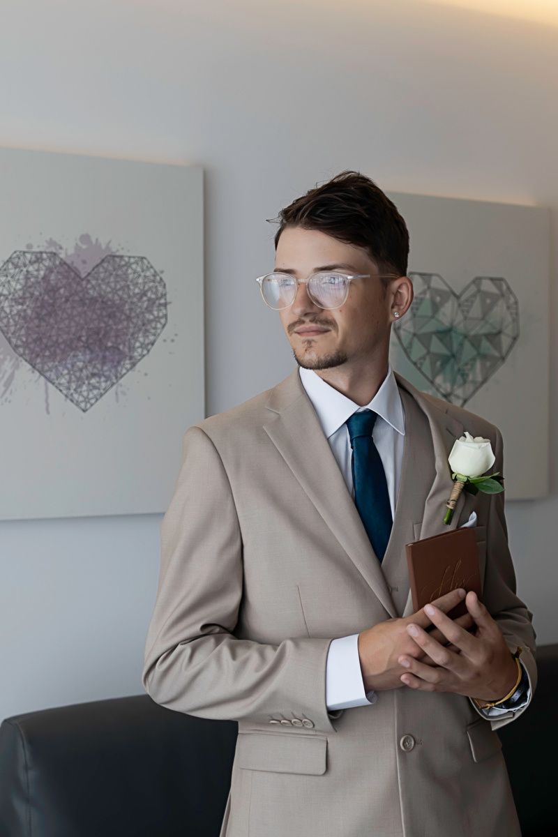 Groom in beige suit with teal tie, white boutonniere, and clear glasses holding a book before wedding ceremony.