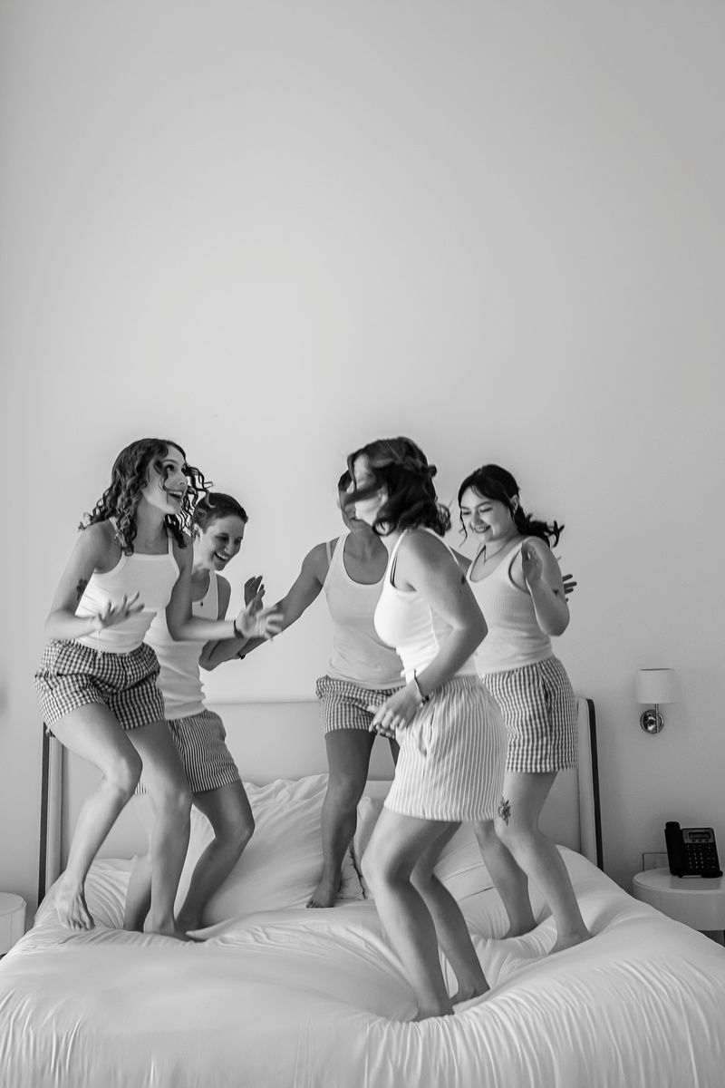 Group of young women dancing and jumping on a white bed in black and white photo.