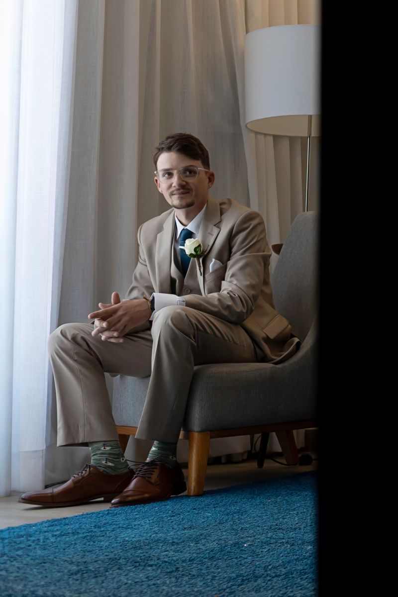 Groom in a tan suit with boutonniere sits smiling in an armchair by curtained window on wedding day.
