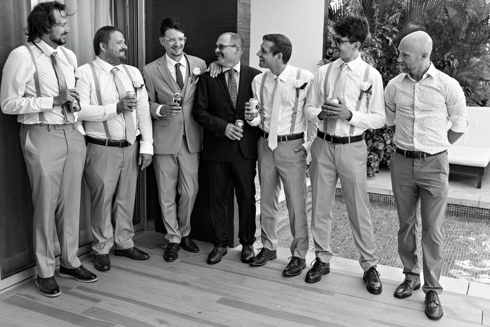 Black and white photo of groom and groomsmen laughing together in suspenders before wedding ceremony.