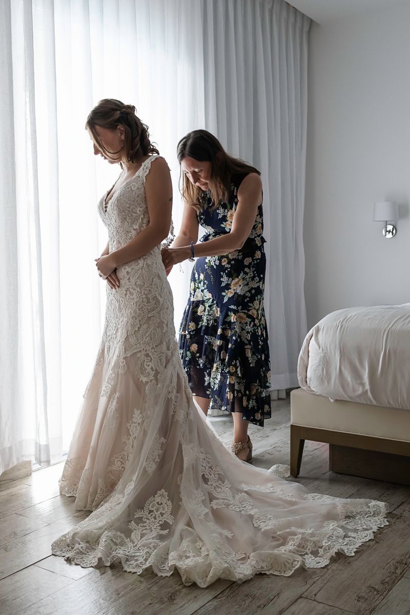 Bride in lace wedding gown being helped by bridesmaid in floral dress near window in hotel room.