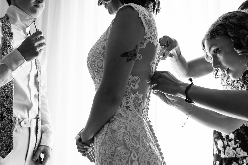 Bridesmaids helping bride button up lace wedding dress in black and white photo.