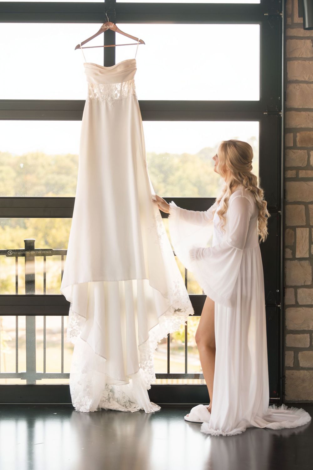 Blonde bride in white robe admires strapless wedding gown hanging by sunlit window.