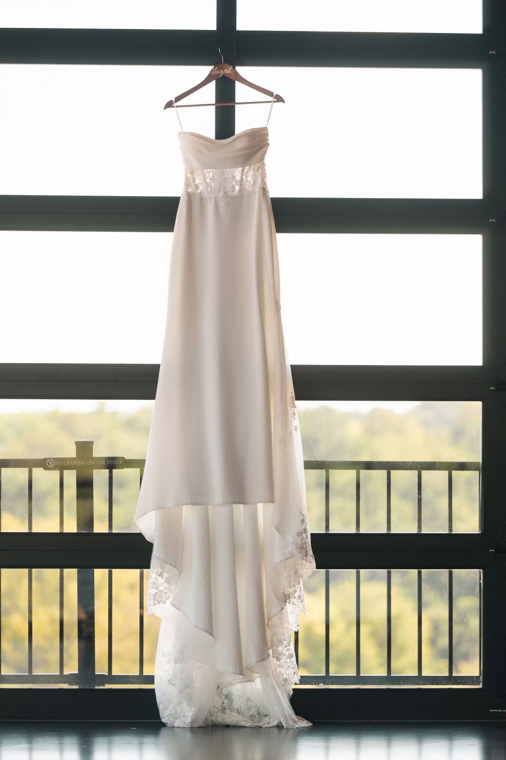 Elegant strapless wedding gown with lace trim hanging on a wooden hanger in front of large windows.