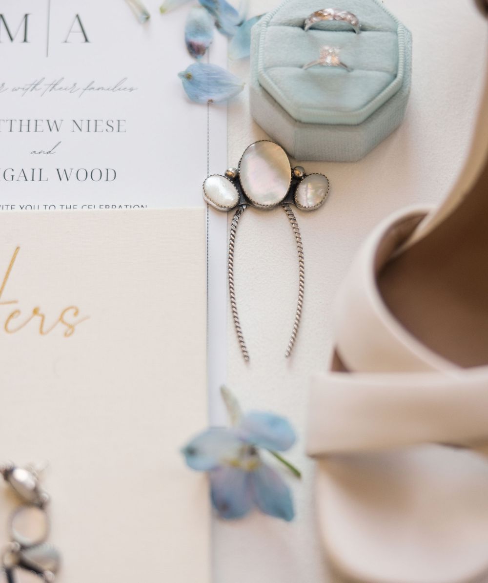 Elegant wedding flat lay with pearl drop earrings, sage velvet ring box, blush heels, and floral invitation suite.