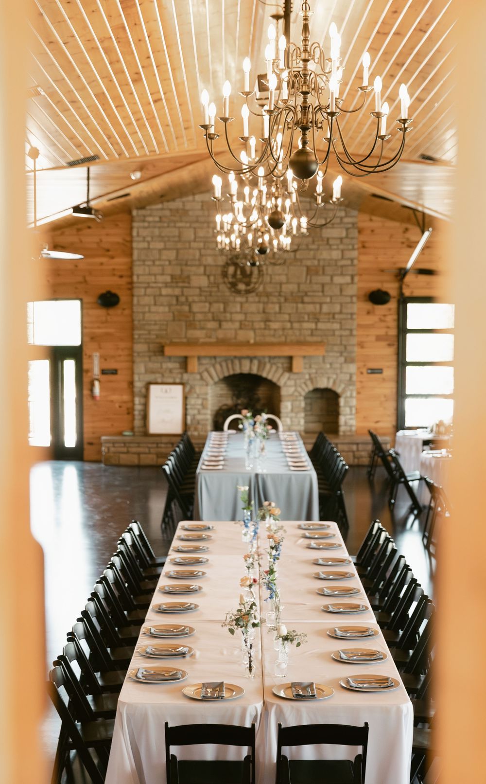 Rustic wedding reception hall with long banquet table, chandelier, and stone fireplace at a barn venue.