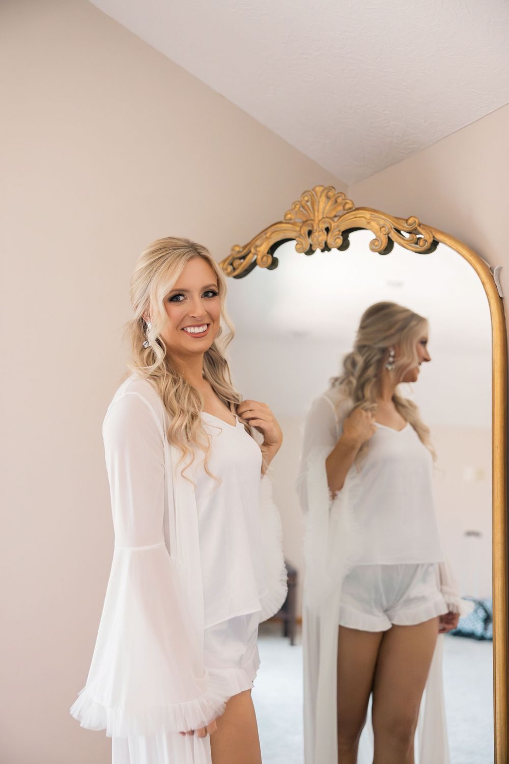 Blonde bride smiles in white bridal robe beside ornate gold mirror during wedding morning preparations.