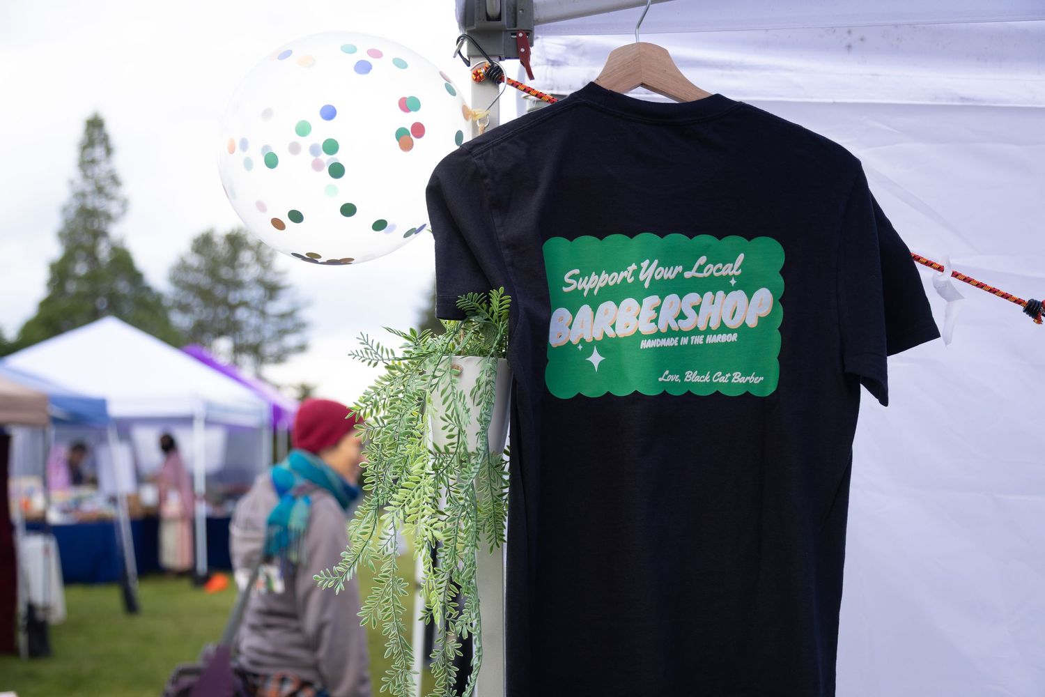 Black t-shirt reading Support Your Local Barbershop hanging at outdoor market booth with balloon and green plants.