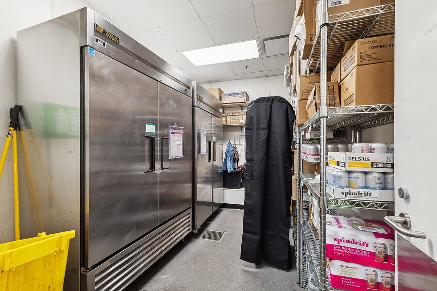 Commercial kitchen storage room with stainless steel refrigerators and wire shelving stocked with supplies.