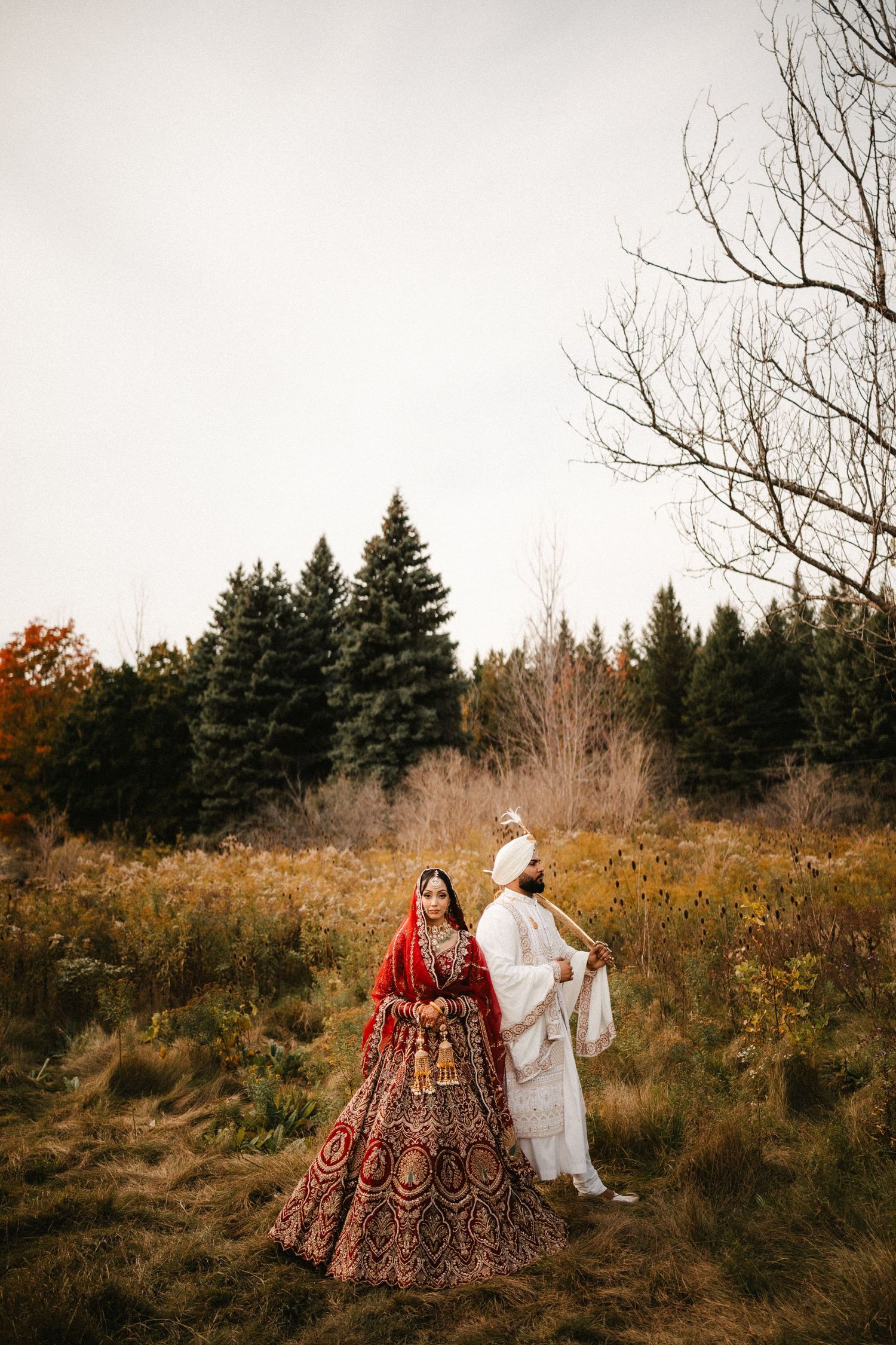 Indian bride in red lehenga and groom in white sherwani stand in autumn field with evergreen trees backdrop.
