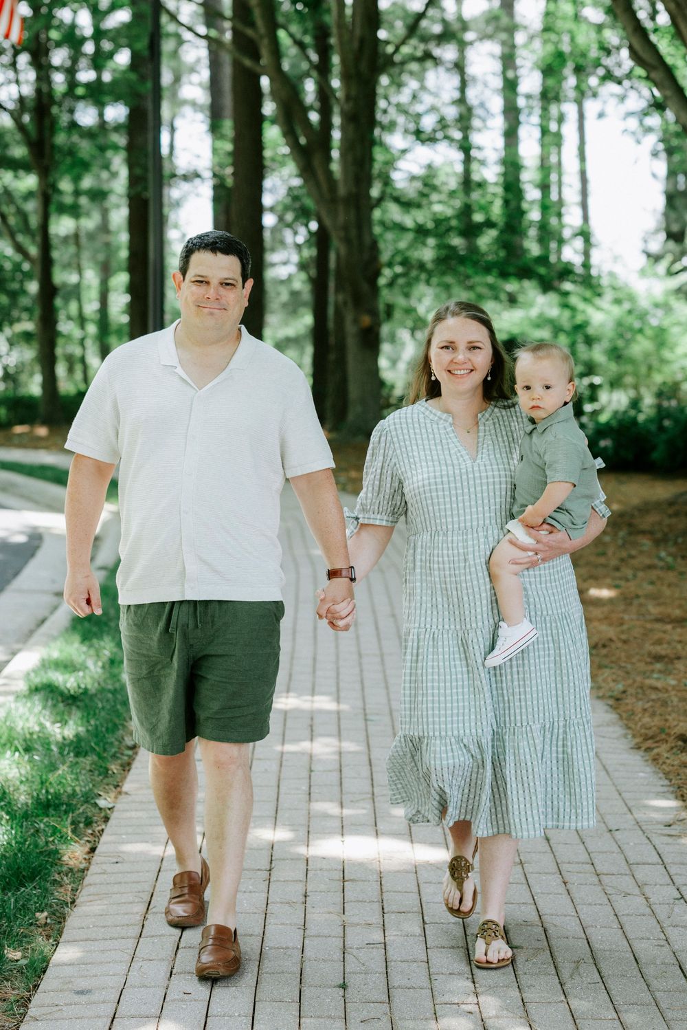 Young family with baby walks hand-in-hand on brick path surrounded by lush green trees on sunny day.