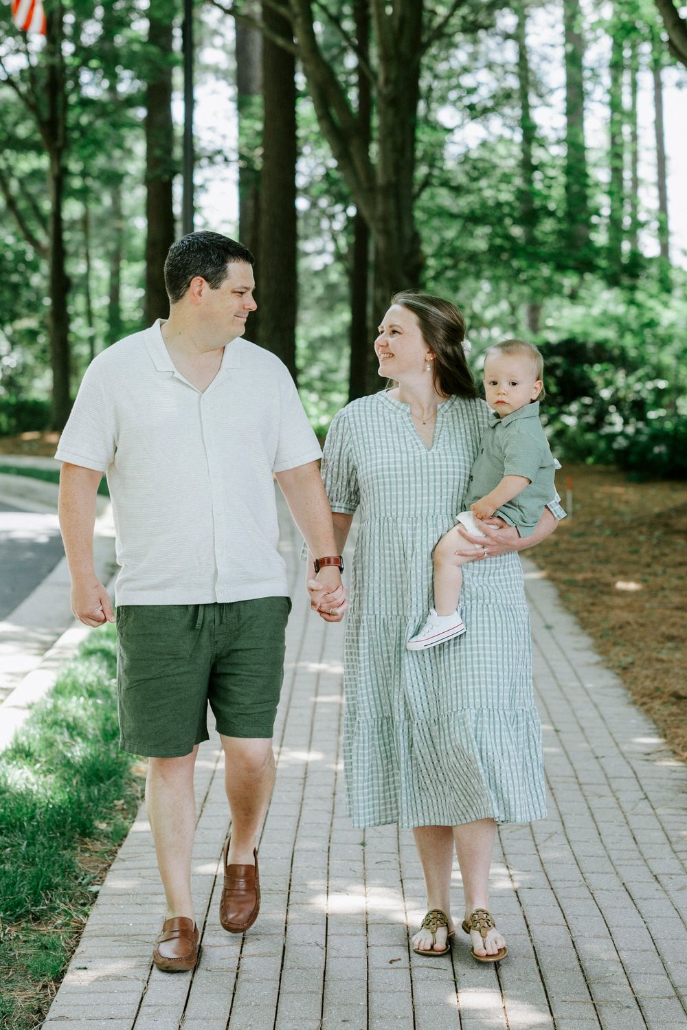 A happy family of three walks hand-in-hand on a brick path surrounded by lush green trees.