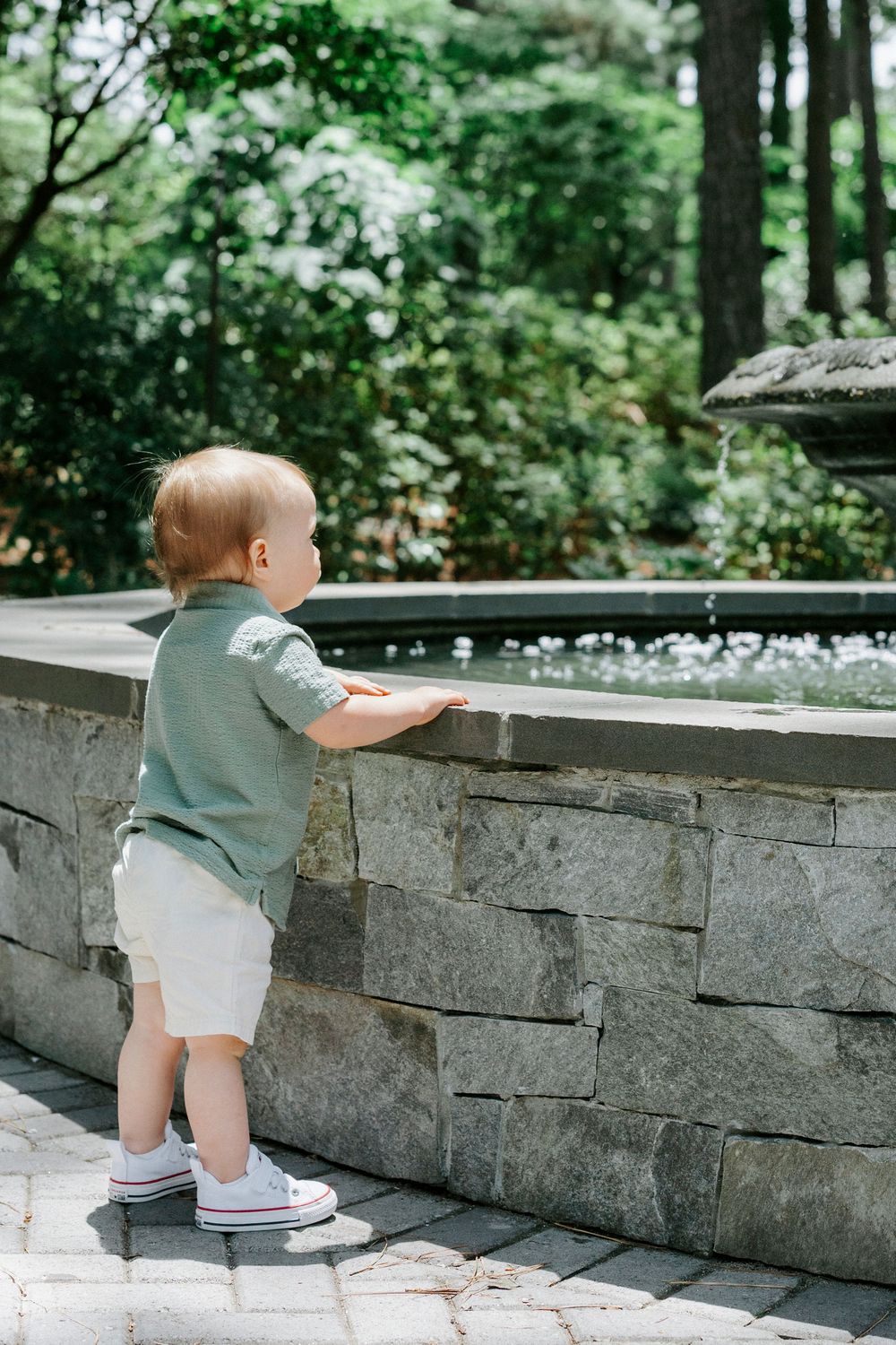 Toddler boy in green shirt and white shorts standing at a stone garden fountain outdoors.