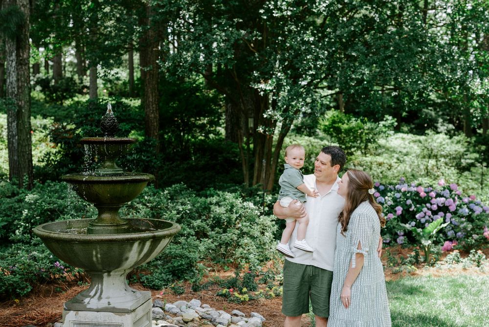 Family with toddler posing near stone fountain in lush green garden with blooming hydrangeas.