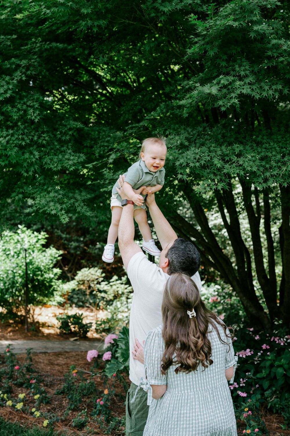 Joyful father tosses laughing baby in air while mother watches in lush green garden setting.