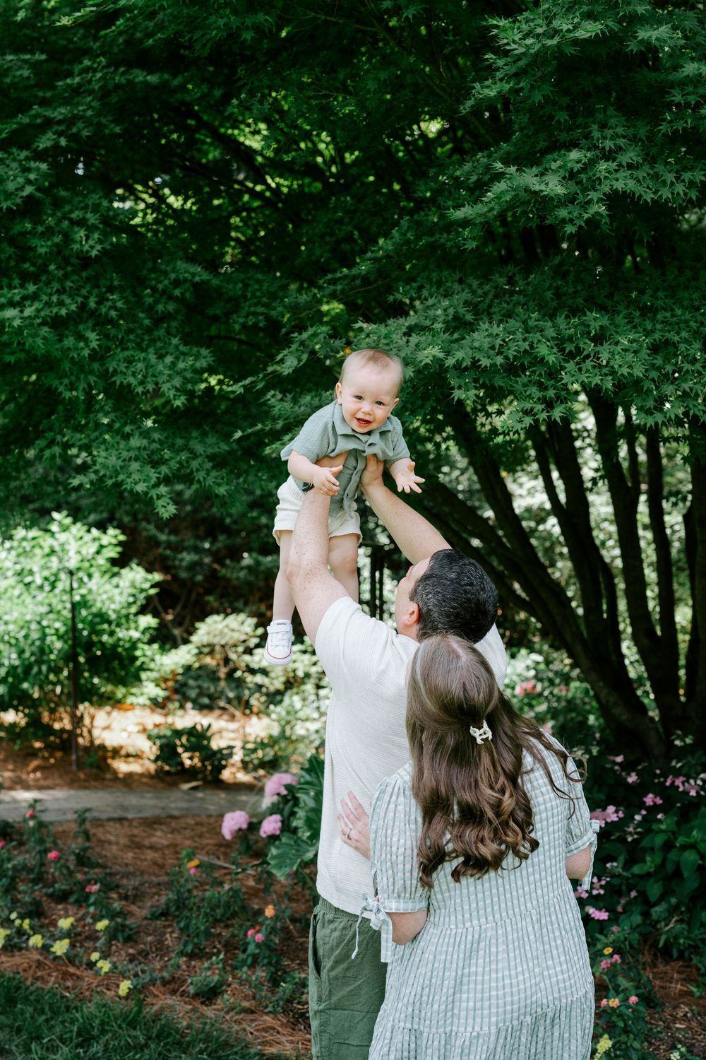 Parents joyfully lifting their baby in a lush green garden with blooming flowers.