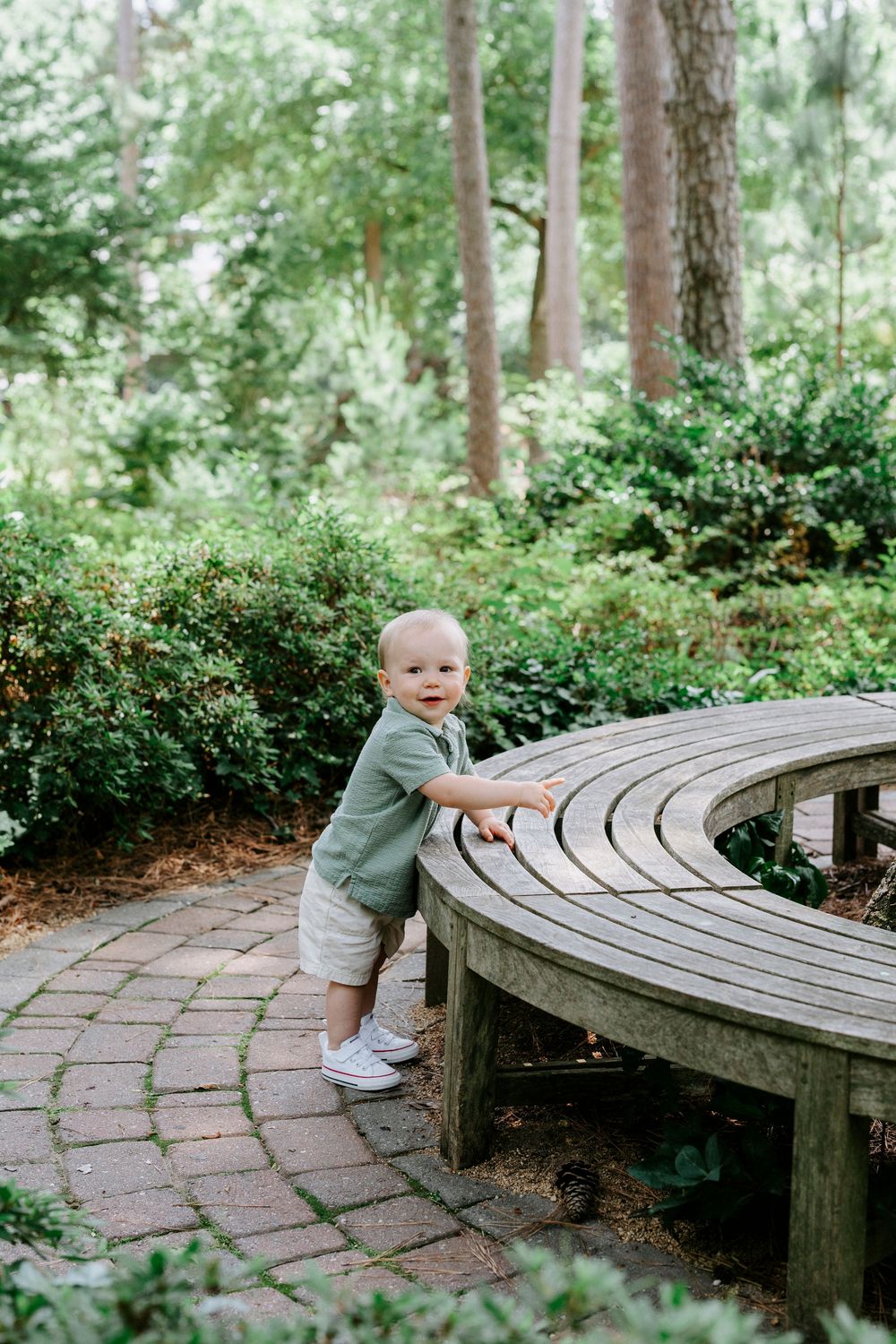 Adorable toddler in green shirt standing by curved wooden bench in garden.