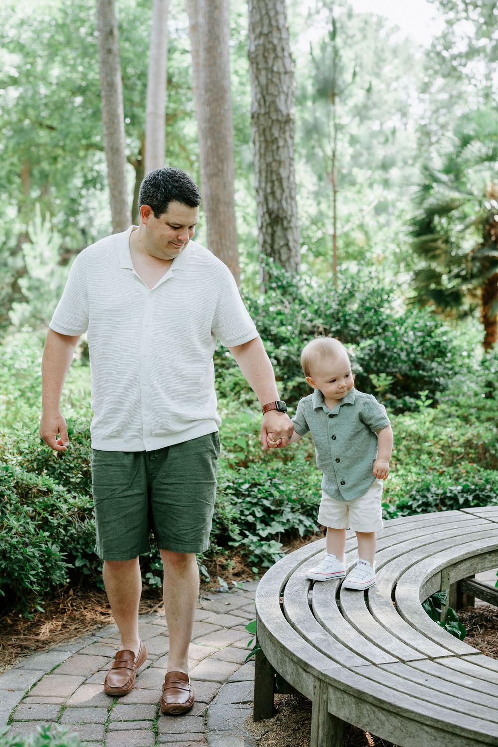 A father holds his toddler's hand on a brick garden path surrounded by lush green trees and shrubs.