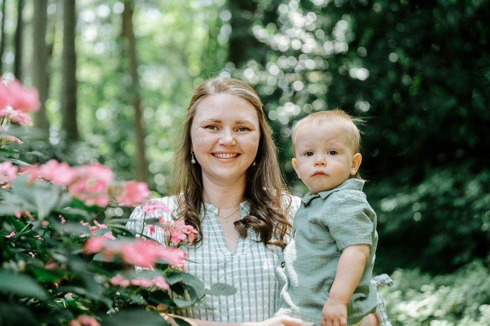 A smiling woman holds a baby boy among pink flowering bushes in a lush green forest garden setting.