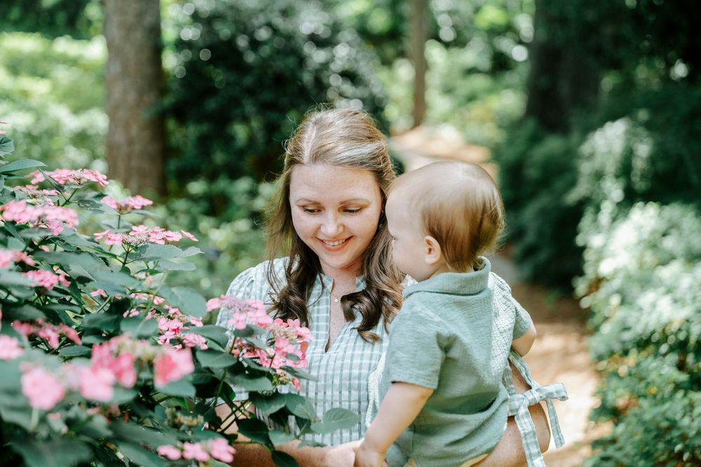 Smiling mother holding baby near pink hydrangeas in a lush garden.