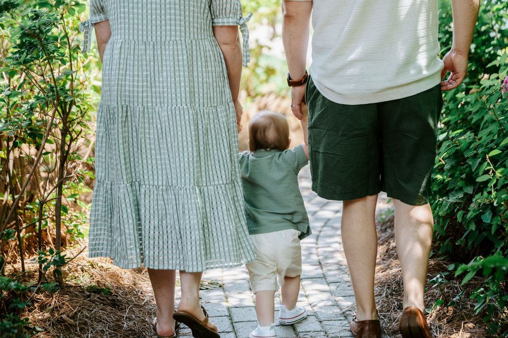 Family of three walking on a garden path, parents holding toddler's hands in lush green surroundings.