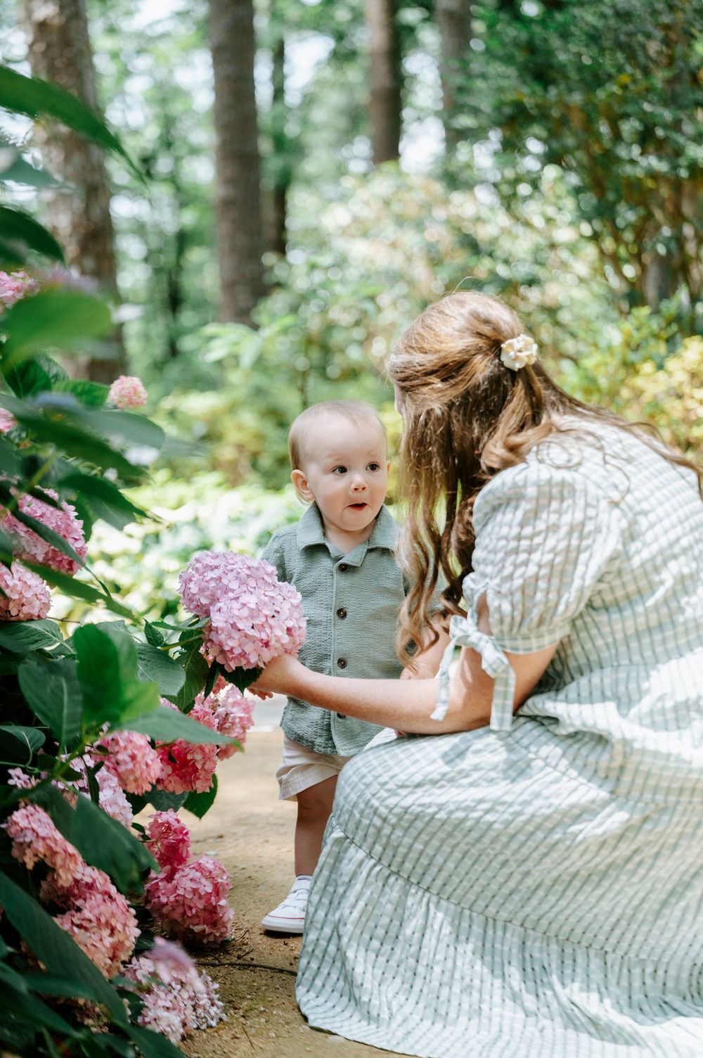Mother and baby boy explore pink hydrangea blooms together in a sunlit garden pathway.
