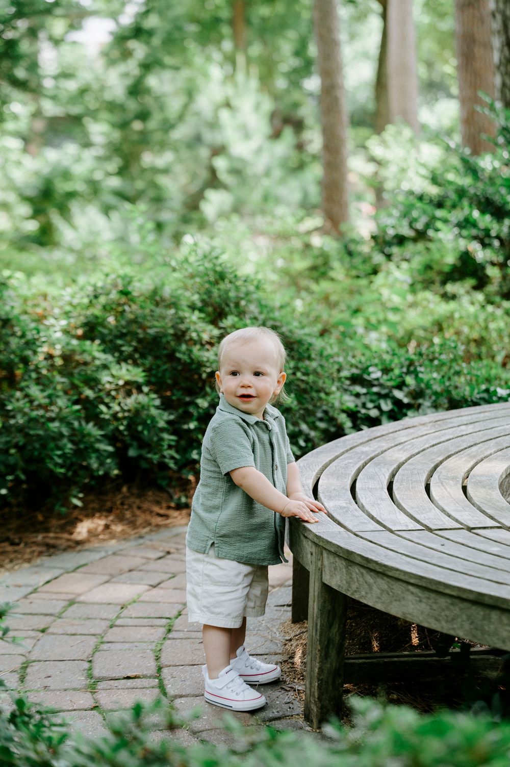 Cute toddler boy in green shirt standing by a circular wooden bench in a garden.