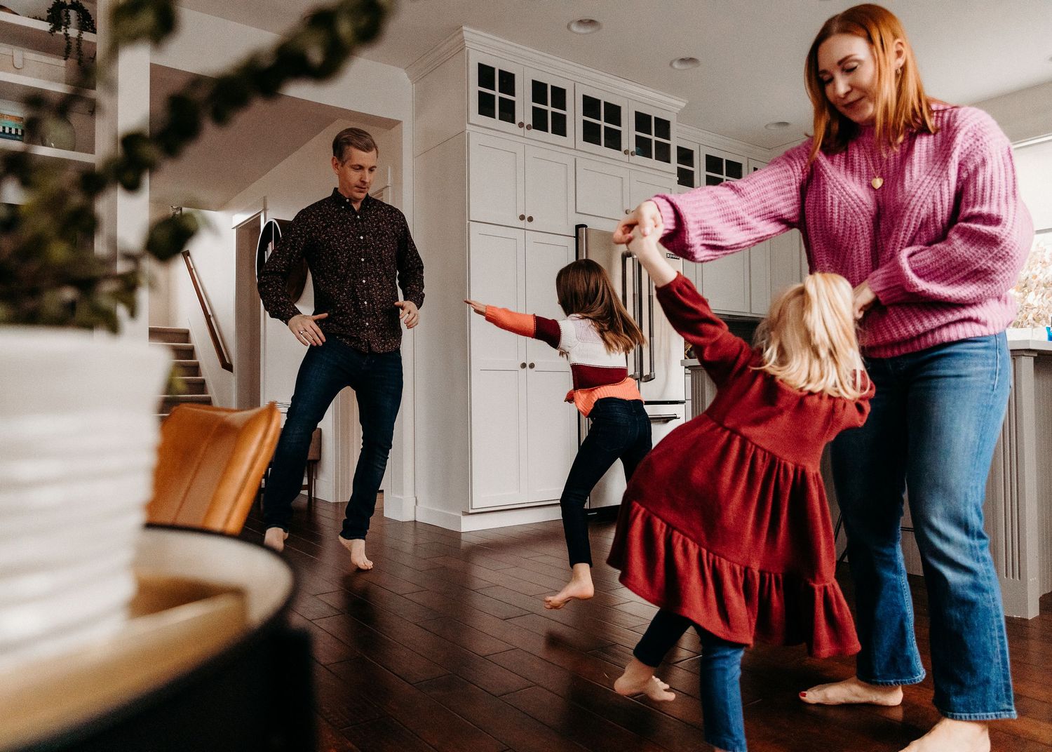 Family dancing together in a bright modern kitchen, parents and two young girls in red dresses spinning joyfully.