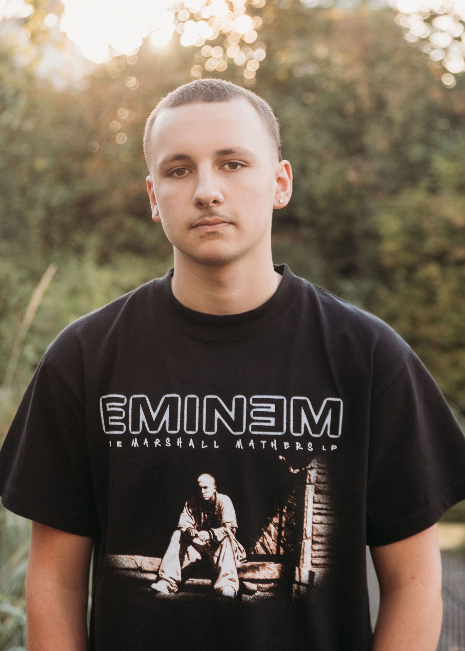 Young man wearing an Eminem Marshall Mathers LP black graphic t-shirt outdoors with green foliage background.