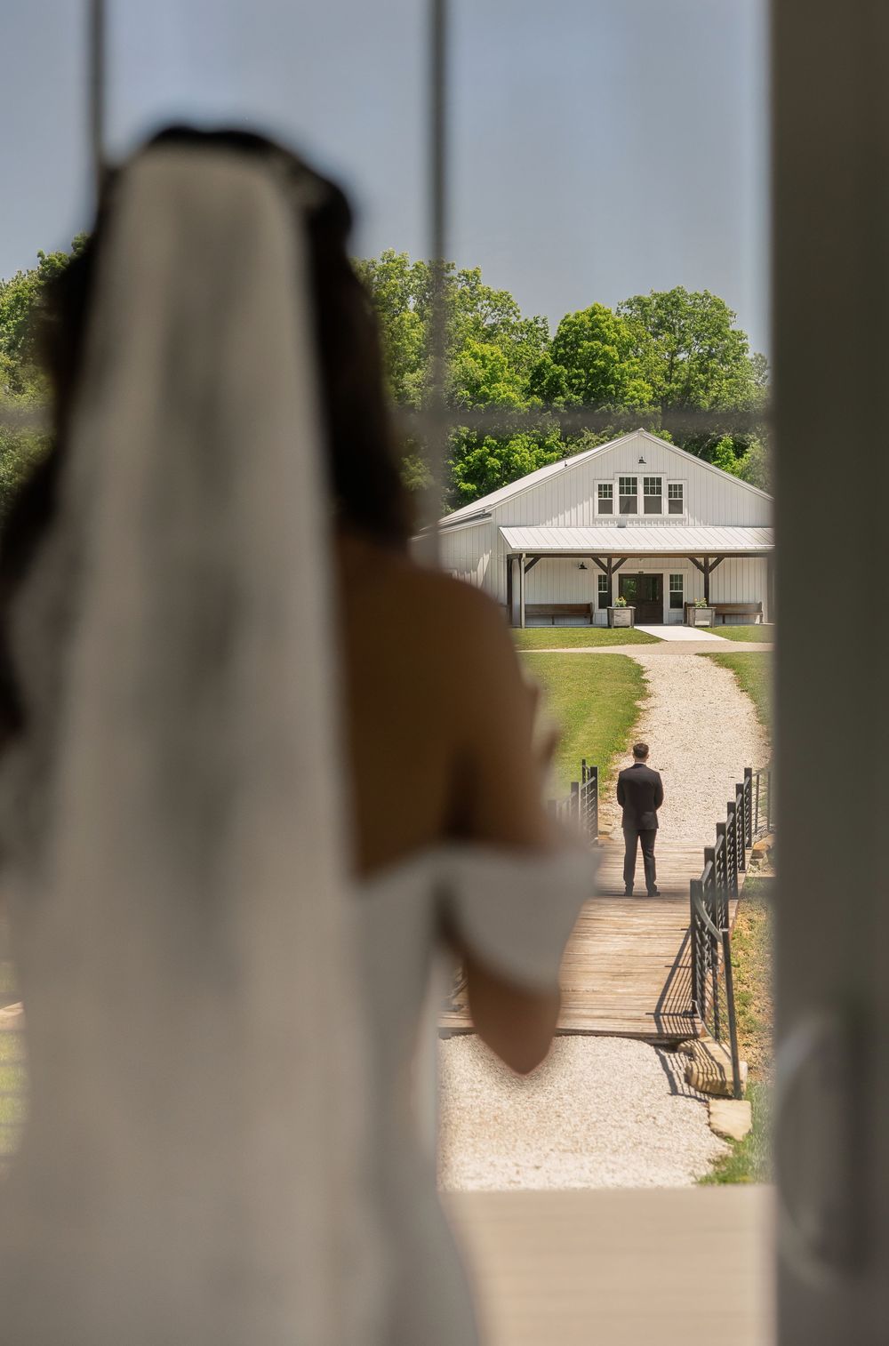 Bride in white gown and veil watches groom standing on path leading to white wedding venue building.