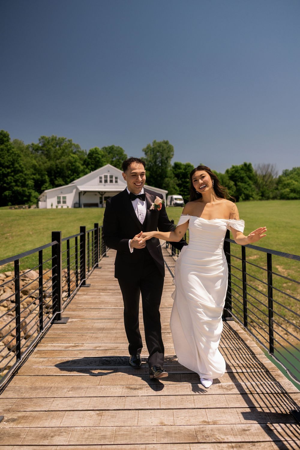 Joyful bride and groom walk hand-in-hand across wooden bridge on sunny wedding day.