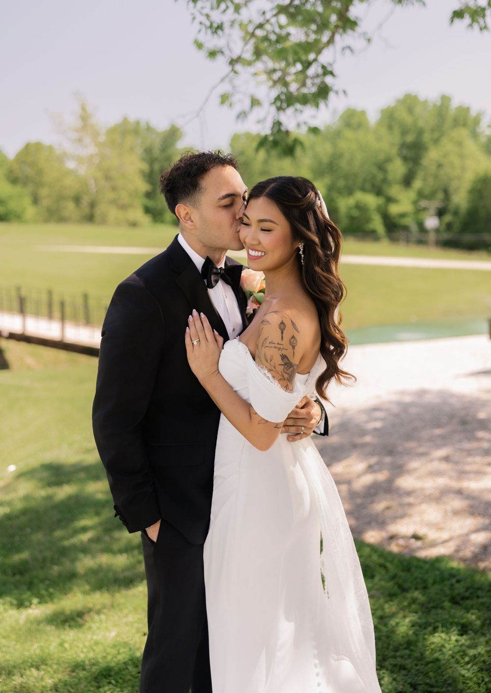 Bride and groom share a romantic moment outdoors, with groom in tuxedo kissing brides cheek near a pond.