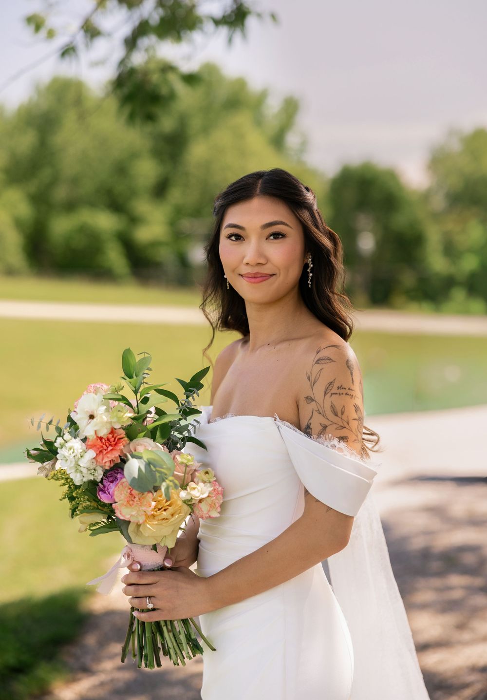 Bride in off-shoulder white gown holds colorful bouquet outdoors, showcasing floral tattoo sleeve and elegant bridal look. williams farm