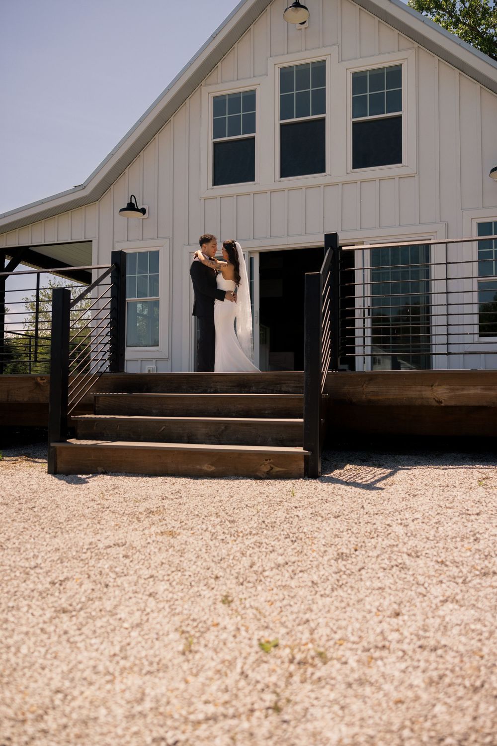Wedding couple shares romantic kiss on steps of white barn venue in sunny outdoor ceremony setting.