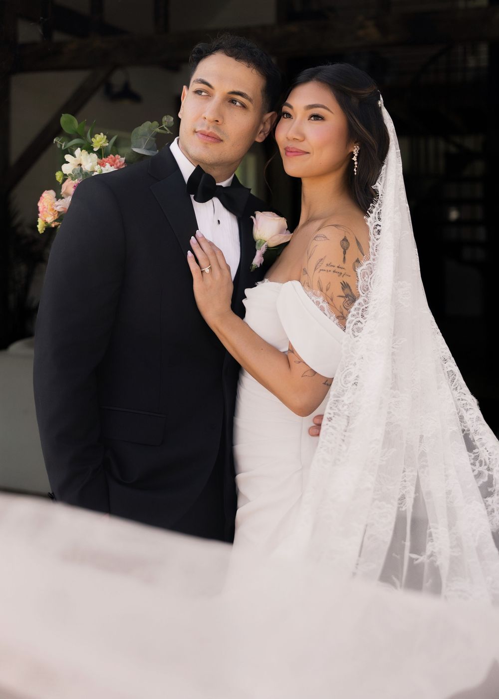 Bride and groom pose together in classic black tuxedo and off-shoulder lace wedding gown with veil.