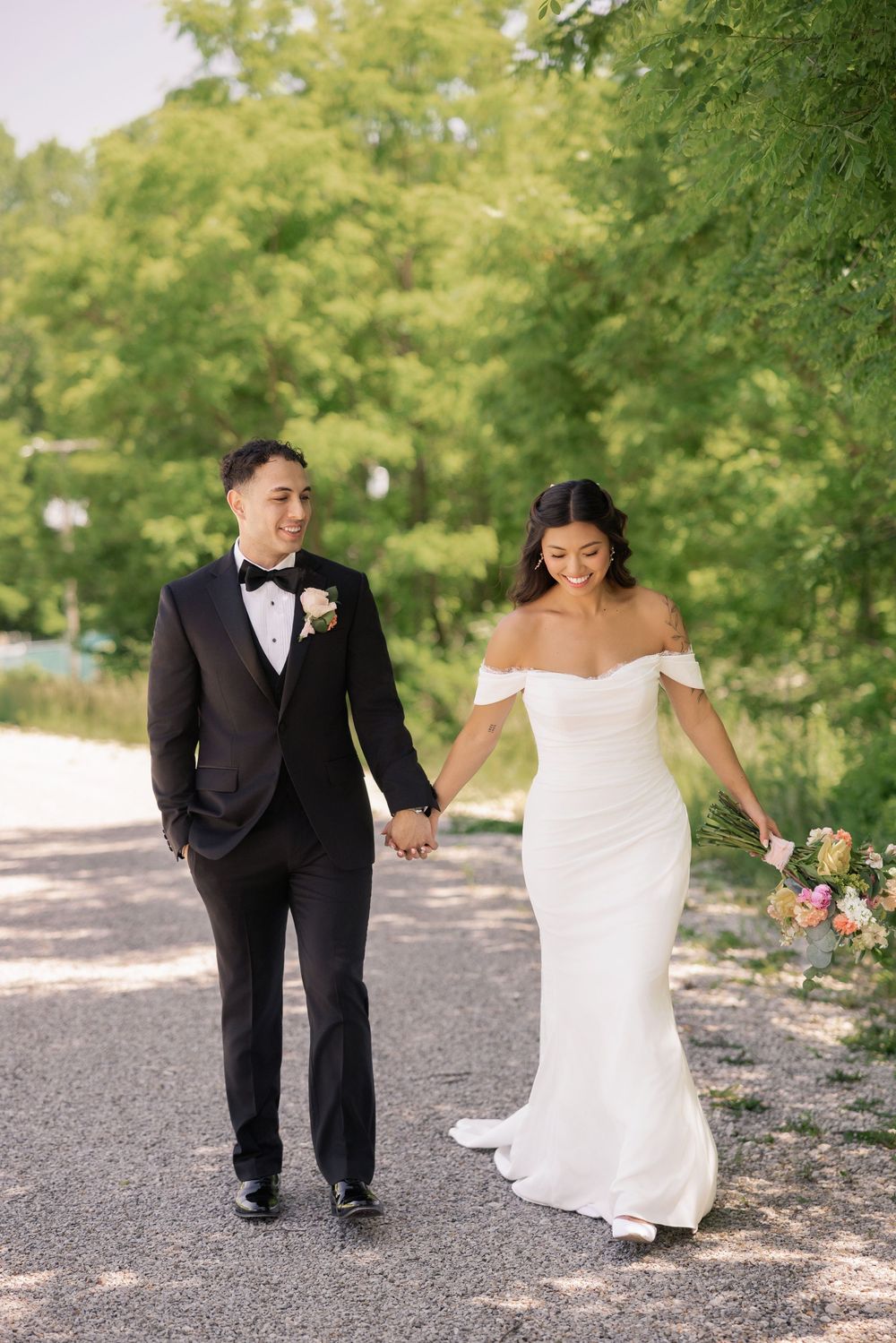 Bride and groom walk hand-in-hand on tree-lined path, bride holding colorful bouquet in white gown.