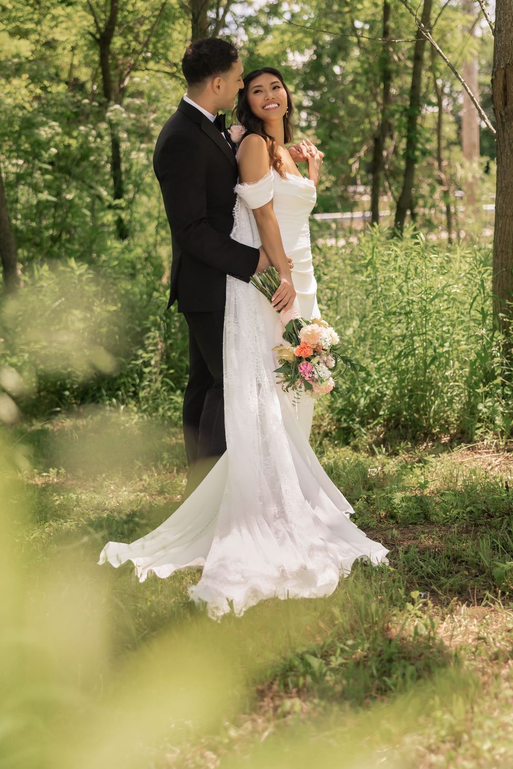 Smiling bride in off-shoulder white gown holds bouquet while groom embraces her in lush green forest. williams farm, pataskala Ohio.