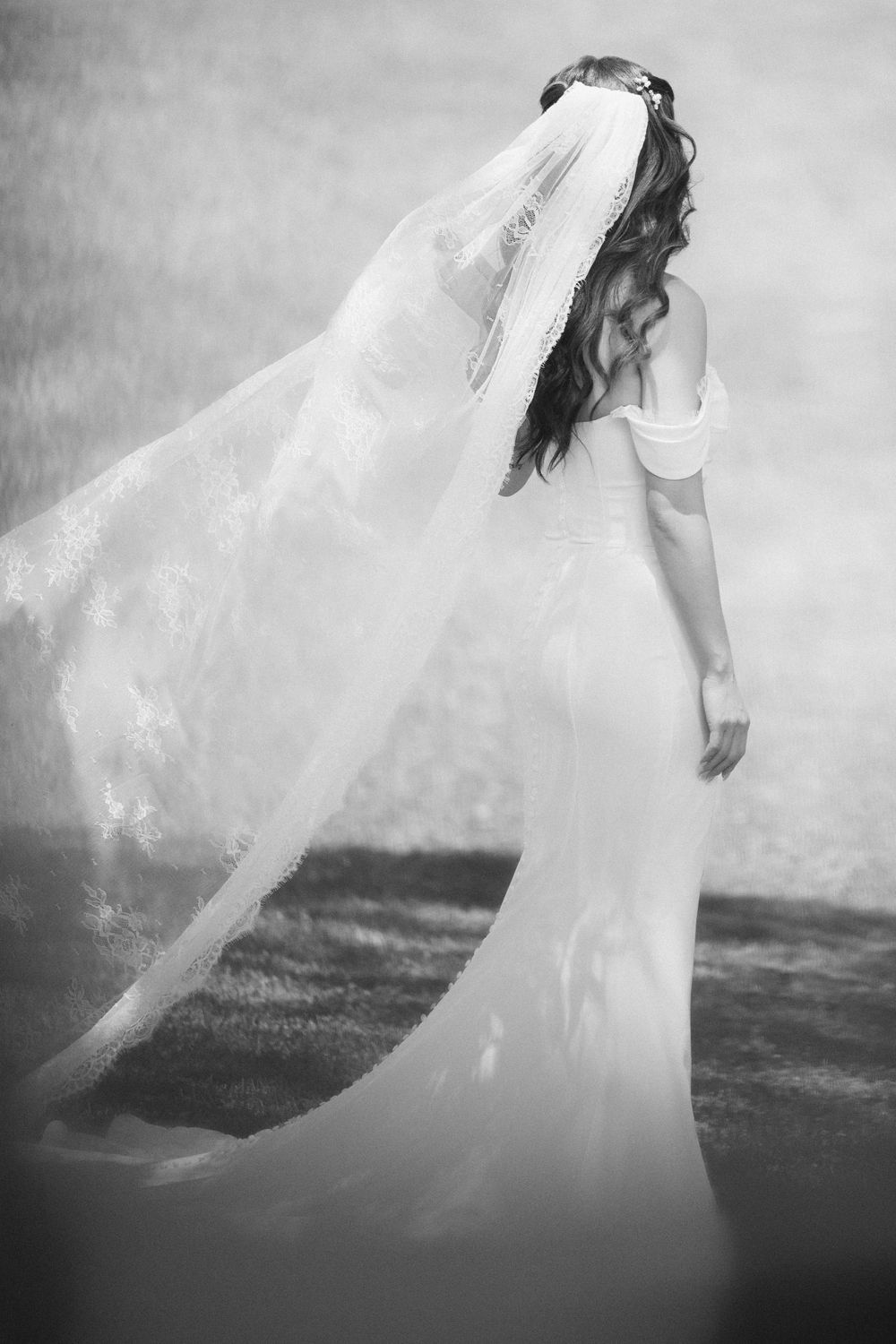 Black and white photo of a bride in a flowing white gown and long lace veil blowing in the wind.