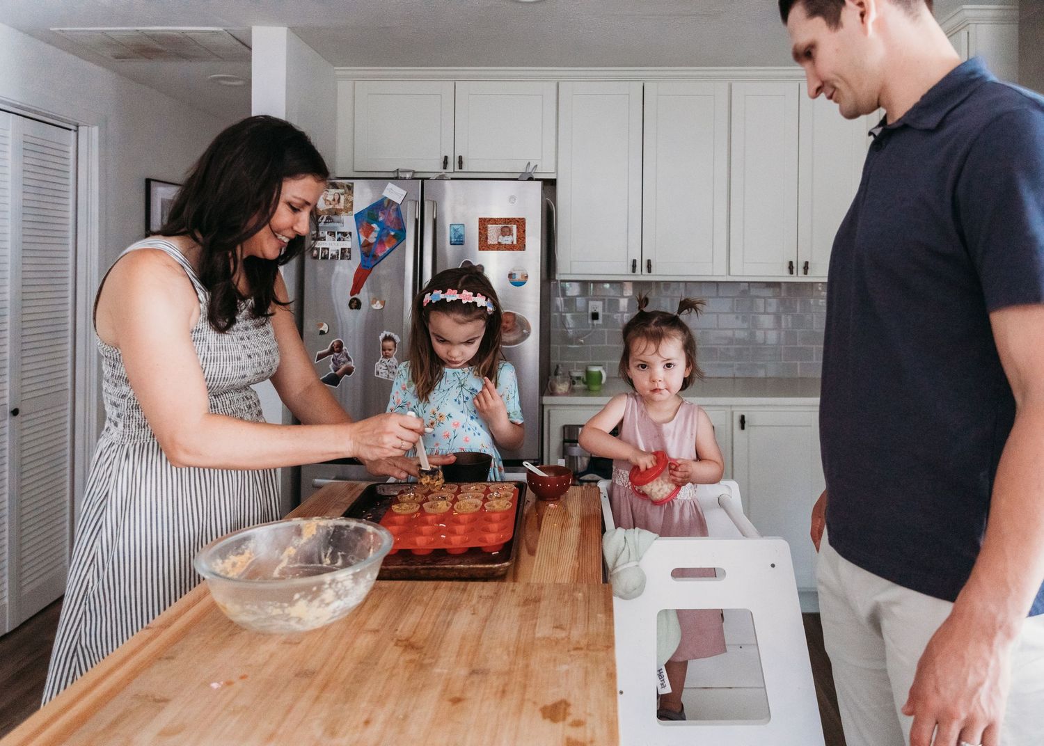 Family baking together in a bright kitchen, parents helping two young girls decorate a muffin tin on a wooden counter.