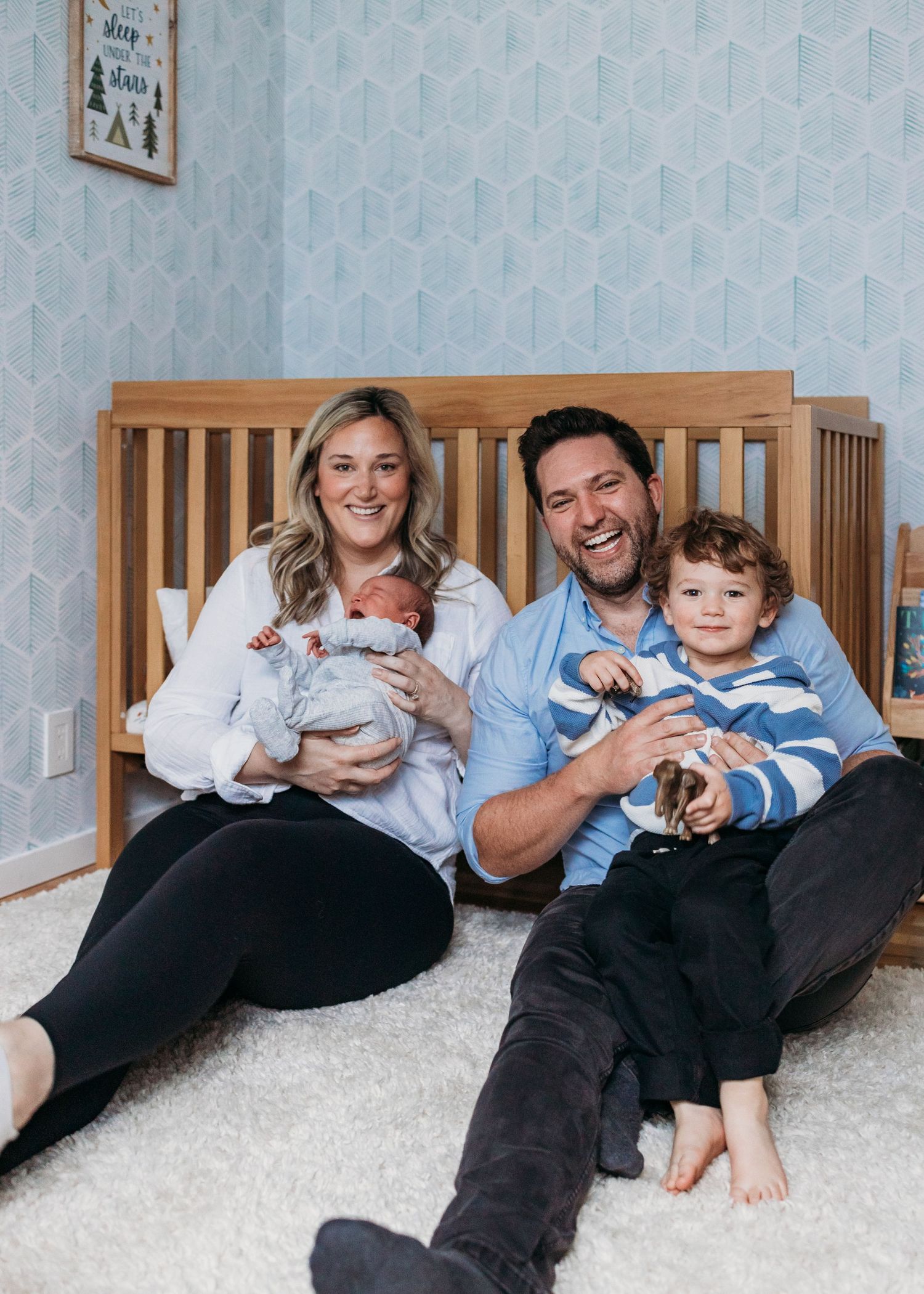 Happy family sitting on nursery floor, parents smiling with newborn baby and toddler son by wooden crib.