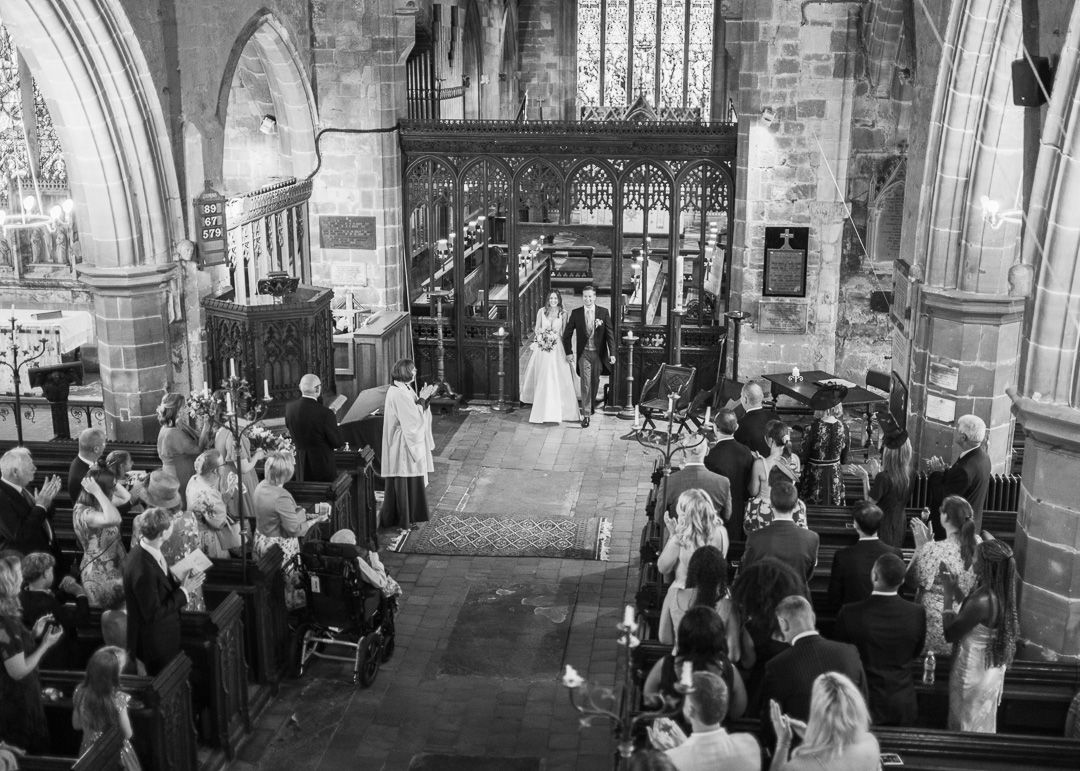 Black and white photo of a traditional church wedding ceremony with guests standing in pews as bride and groom exchange vows.