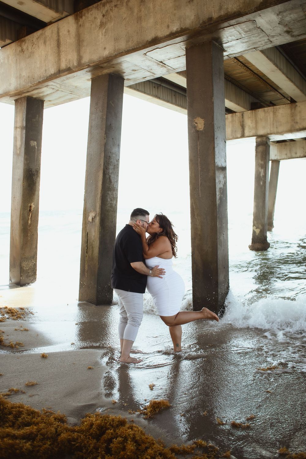 Couple kissing under Pompano Beach Pier during engagement couples session.