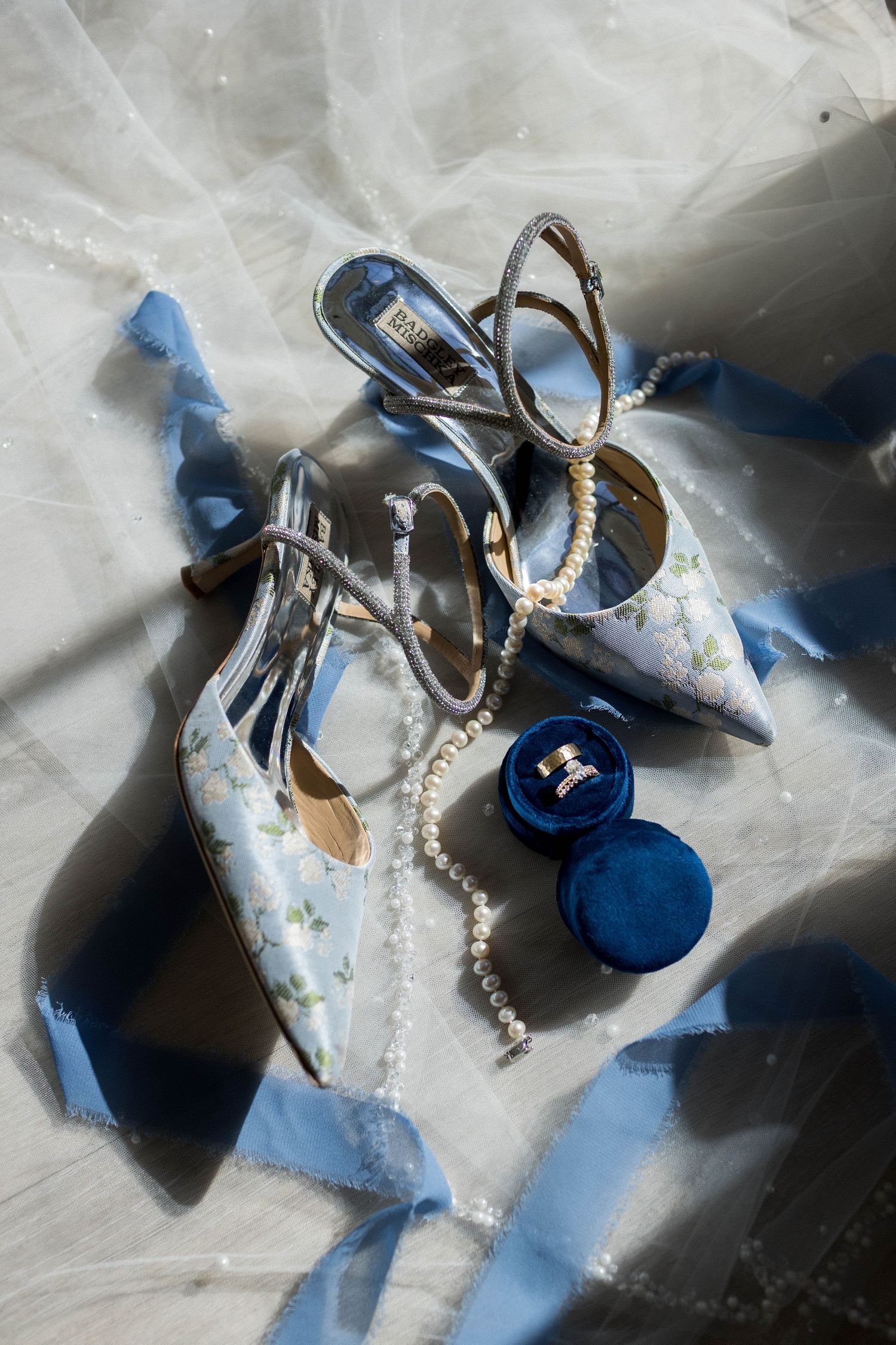 Elegant bridal accessories featuring floral heels, pearl necklace, gold rings in blue velvet box, and blue ribbon on white veil.