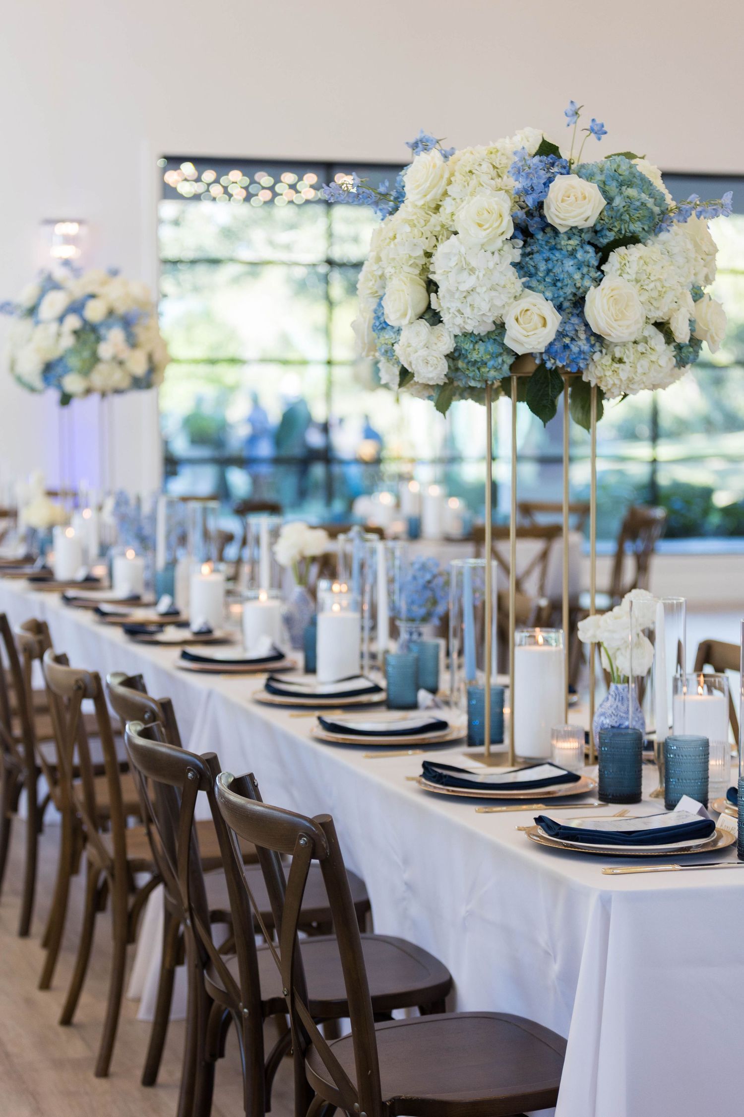 Elegant wedding reception table with white and blue hydrangea centerpieces, white linens, and dark wood cross-back chairs.