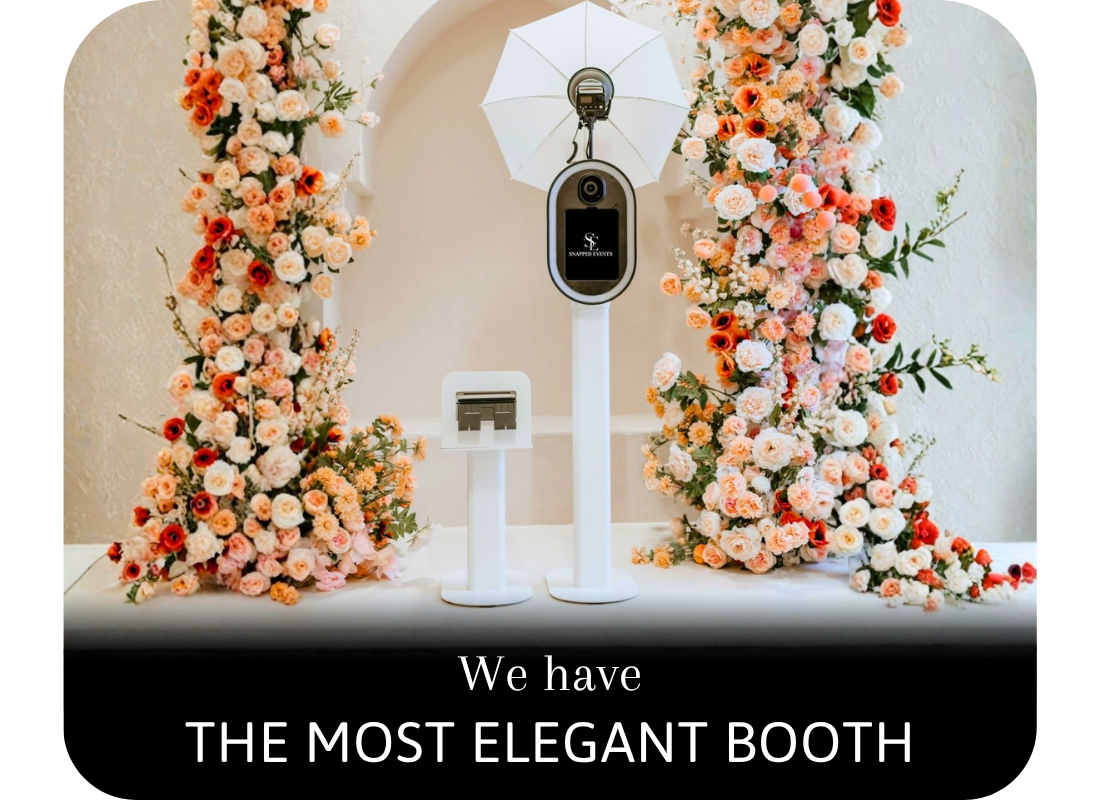 Elegant photo booth stand with umbrella light surrounded by lush pink and white floral arrangements at a wedding venue.