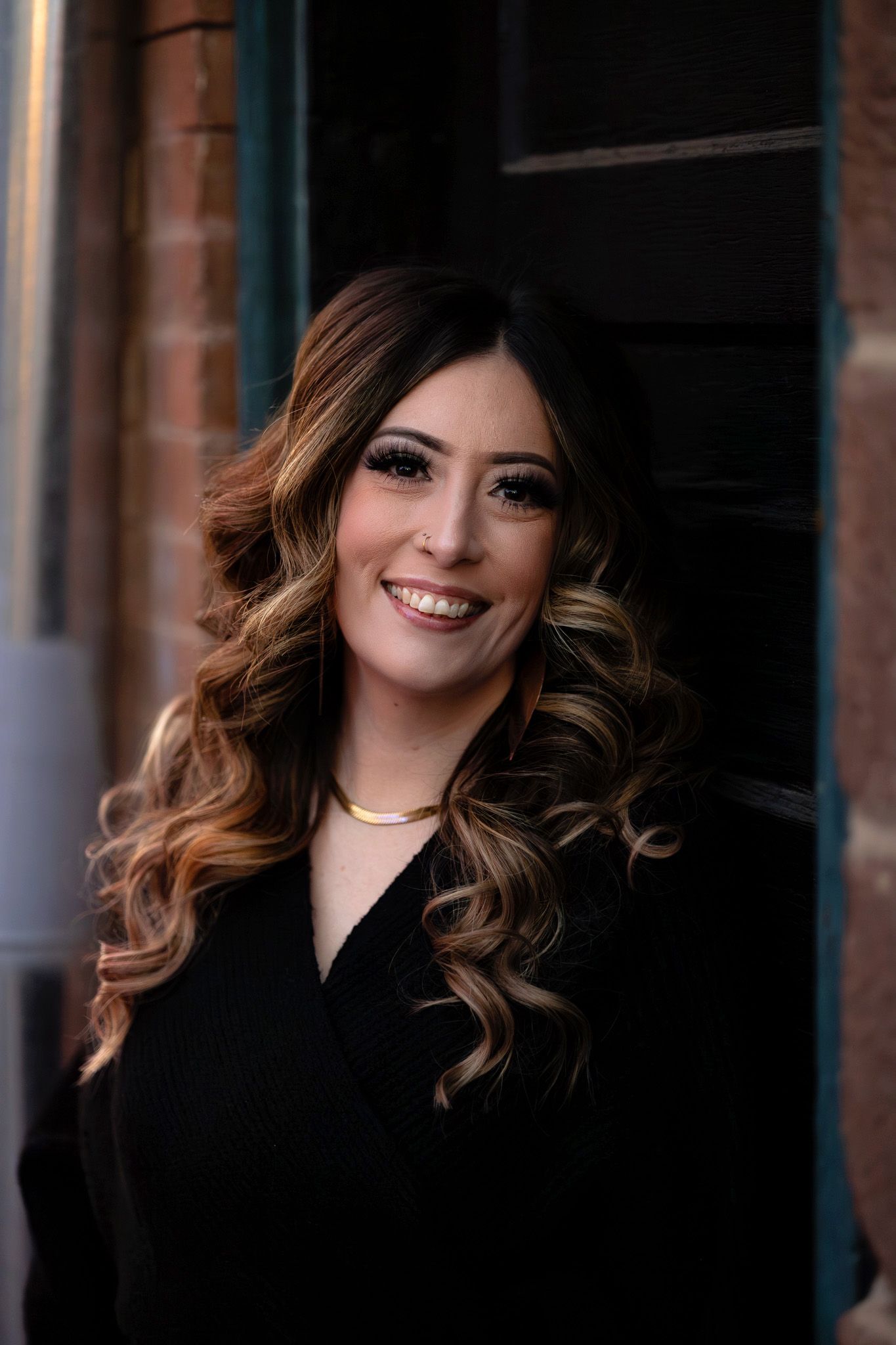 A smiling woman with curly highlighted hair wearing a black top and gold necklace poses outdoors near a brick wall.